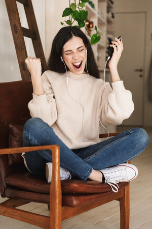 Photo of excited caucasian woman 20s wearing earphones singing and holding cell phone while listening to music indoorの写真素材