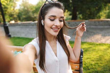 Beautiful young woman taking a selfie while sitting on a bench with shopping bags outdoorsの写真素材