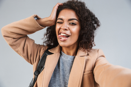 Cheerful young african woman wearing coat standing isolated over gray background, taking a selfieの写真素材