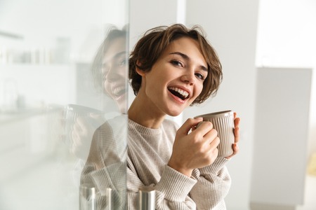 Side view of Happy brunette woman drinking coffee and looking at the camera at home