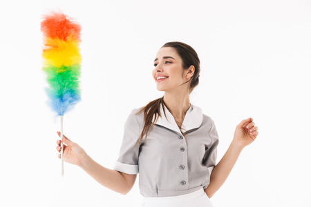 Photo of adorable maid 20s dressed in uniform holding colorful duster while doing housework and...