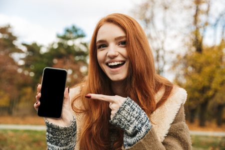 Lovely redheaded young girl listening to music while sitting on a bench, using mobile phone, showing blank screen mobile phoneの写真素材