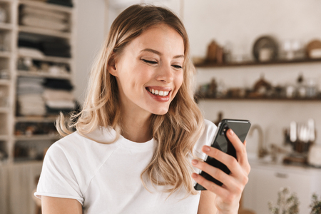 Portrait of european blond woman 20s wearing casual t-shirt using smartphone while standing in stylish wooden kitchen at homeの写真素材