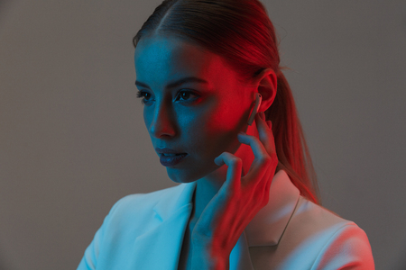 Photo closeup of charming woman 20s wearing earpods looking at camera while standing under neon lights isolated over gray backgroundの写真素材