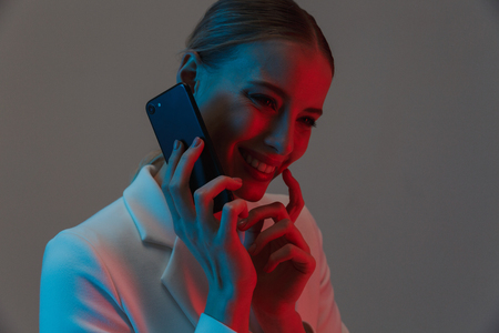 Image closeup of elegant woman 20s with ponytail hairstyle holding and talking on mobile phone while standing under neon lights isolated over gray backgroundの写真素材