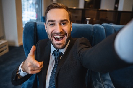 Handsome young excited businessman wearing suit sitting at the hotel lobby, using mobile phone, taking a selfie, pointingの写真素材