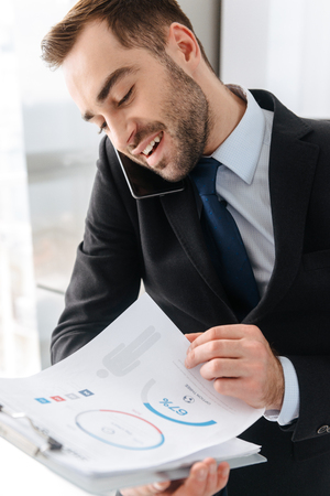 Image of pleased businessman 30s in formal suit talking on mobile phone while holding clipboard with paper charts in bright apartmentの写真素材
