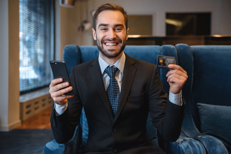 Handsome young businessman wearing suit sitting at the hotel lobby, using mobile phone, showing plastic credit cardの写真素材