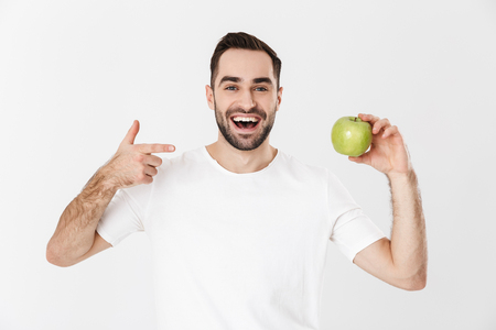 Handsome cheerful man wearing blank t-shirt standing isolated over white background, showing green apple, pointingの写真素材