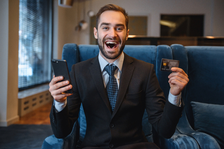 Handsome young businessman wearing suit sitting at the hotel lobby, using mobile phone, showing plastic credit cardの写真素材