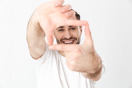 Handsome cheerful man wearing blank t-shirt standing isolated over white background, showing photographing gestureの写真素材