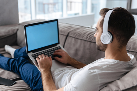 Photo of modern guy 30s in casual t-shirt wearing headphones holding and using silver laptop while lying on sofa in bright apartmentの写真素材