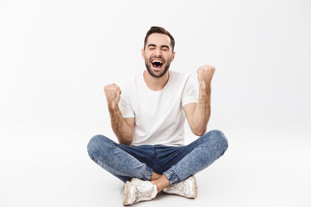 Full length of a cheerful young man sitting with legs crossed isolated over white background, celebrating successの写真素材