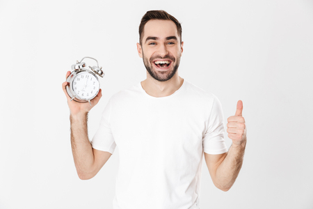 Handsome cheerful man wearing blank t-shirt standing isolated over white background, showing alarm clockの写真素材