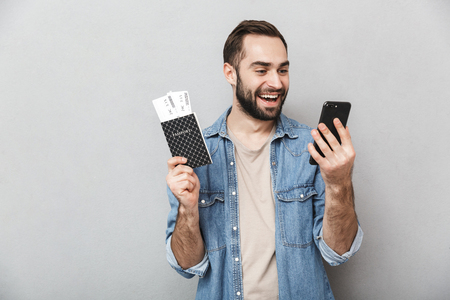 Excited cheerful man wearing shirt isolated over gray background, holding passport with flight tickets, using mobile phoneの写真素材