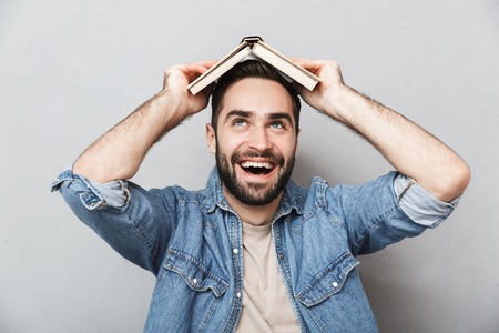 Excited cheerful man wearing shirt isolated over gray background, holding open book on his head