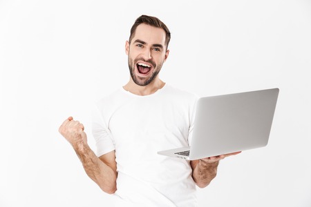 Handsome cheerful man wearing blank t-shirt standing isolated over white background, using laptop computer, celebrting successの写真素材