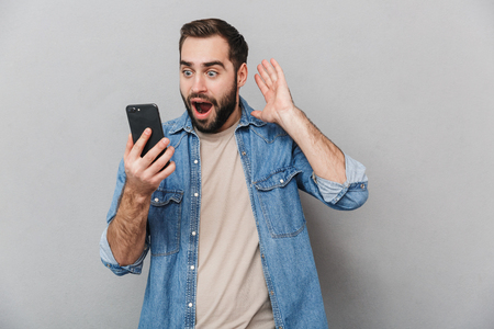 Excited cheerful man wearing shirt isolated over gray background, using mobile phoneの写真素材
