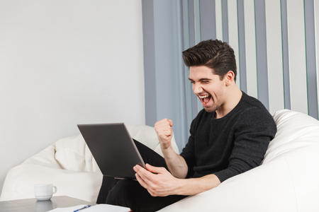 Image of a happy handsome young man at home indoors using laptop computer make winner gesture.の写真素材