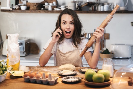 Cheerful young woman wearing apron preparing dough for an apple pie at the kitchen at home, using mobile phone, holding rolling pinの写真素材