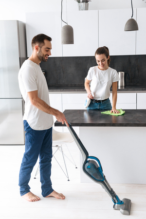 Happy young couple doing general clean at the kitchen, man vacuuming the floor, woman cleaning kitchen deskの写真素材