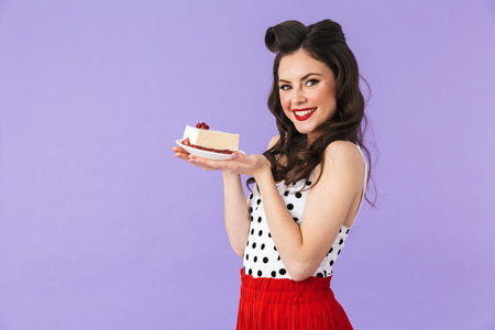 Portrait of positive pin-up woman 20s in vintage polka dot dress smiling while holding and eating sweet cheesecake isolated over violet backgroundの写真素材
