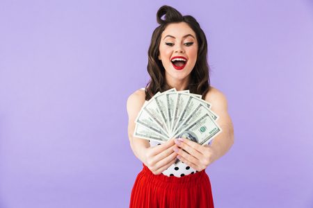 Portrait of a beautiful pin-up girl wearing bright makeup standing isolated over violet background, showing money banknotesの写真素材