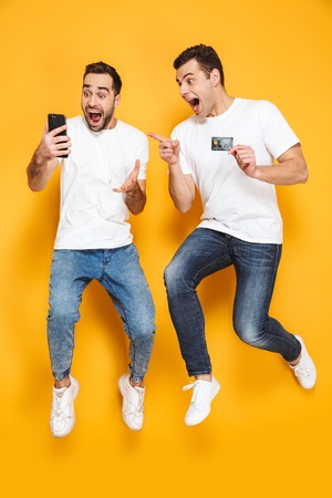 Full length of two cheerful excited men friends wearing blank t-shirts jumping isolated over yellow background, looking at mobile phone, celebrating successの写真素材