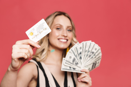Photo of a beautiful amazing young woman posing isolated over red coral background holding credit card and money.の写真素材