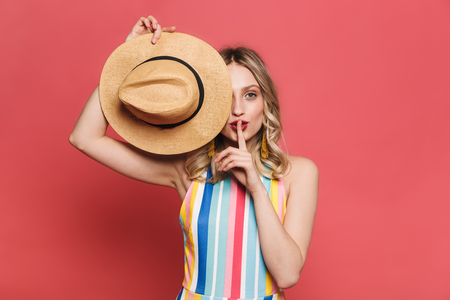 Image of a beautiful amazing young woman posing isolated over red coral background holding hat showing silence gesture.の写真素材