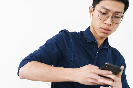 Photo of attractive chinese businessman 20s wearing earpods sitting in chair and holding cell phone while working in office isolated over white backgroundの写真素材