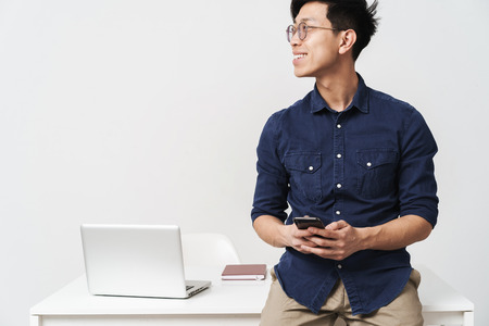 Photo of successful asian businessman 20s wearing eyeglasses sitting at table and holding smartphone while working with laptop in office isolated over white backgroundの写真素材