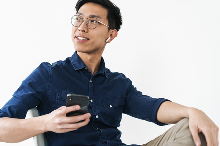 Photo of cheerful chinese businessman 20s wearing earpods sitting in chair and holding cell phone...
