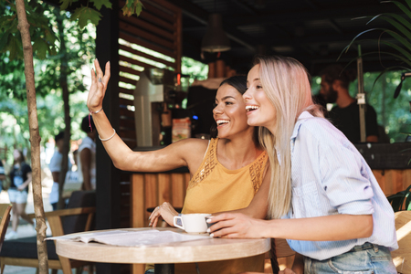 Two cheerful young girlfriends sitting at the cafe table indoors, waving handの写真素材