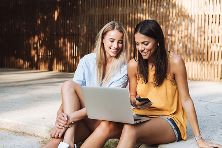 Two smiling young girls using laptop at the park, laughing, chattingの写真素材