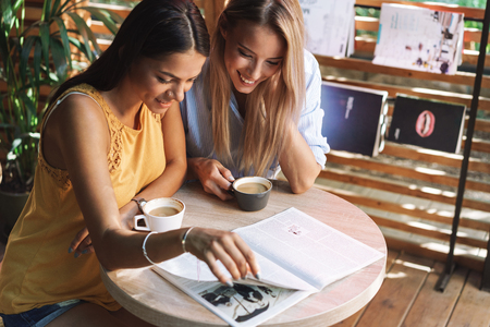 Two cheerful young girlfriends sitting at the cafe indoors, drinking coffee, reading magazineの写真素材