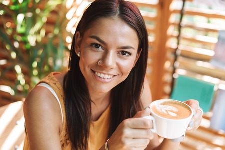 Smiling young woman sitting at the cafe indoors, drinking coffeeの写真素材