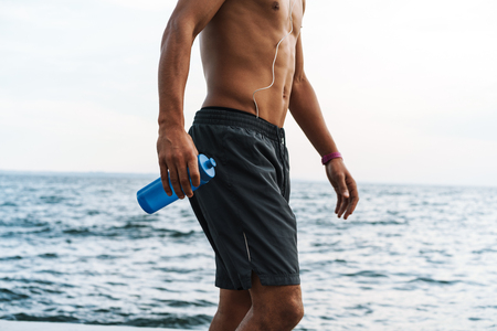 Cropped image of a strong handsome young african sports man outdoors at the beach sea holing bottle with water.の写真素材