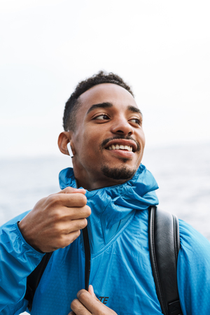 Picture of a handsome happy young african sports man outdoors at the beach sea walking with bag listening music with earphones.の写真素材