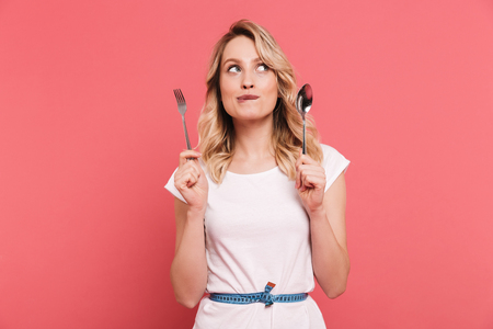 Portrait of thoughtful blond woman 20s wearing body measuring tape around waist holding spoon and fork isolated over pink backgroundの写真素材