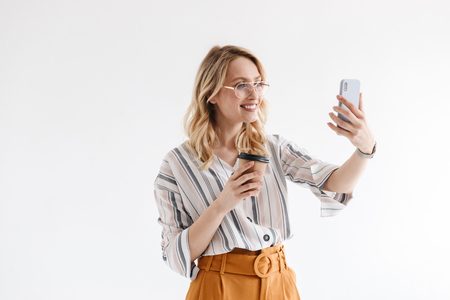Photo of smiling blonde woman wearing glasses taking selfie photo and holding paper cup isolated over white background in studioの写真素材