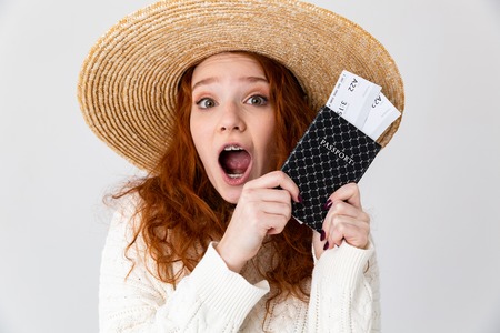 Close up portrait of a cheerful young teenage girl wearing summer hat standing isolated over white background, showing passport with flight ticketsの写真素材