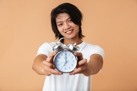 Photo closeup of asian guy 20s wearing white t-shirt smiling and holding alarm clock while looking at camera isolated over beige backgroundの写真素材