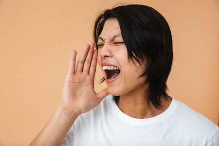 Photo closeup of outraged asian guy 20s wearing white t-shirt frowning and screaming isolated over beige backgroundの写真素材