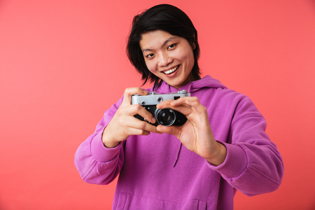 Portrait of a cheerful asian man standing isolated over pink background, taking a picture with photo cameraの写真素材