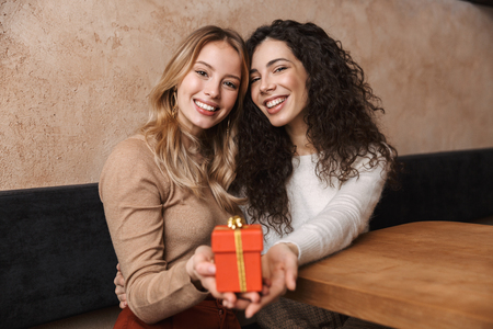 Image of excited happy pretty girls friends sitting in cafe holding present gift box.の写真素材