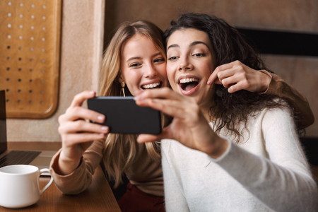 Image of excited happy pretty girls friends sitting in cafe using mobile phone.の写真素材