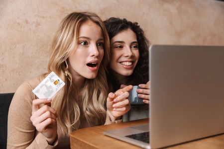 Image of excited shocked pretty girls friends sitting in cafe using laptop computer holding credit card.の写真素材