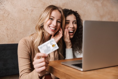 Image of excited pretty girls friends sitting in cafe using laptop computer holding credit card.の写真素材