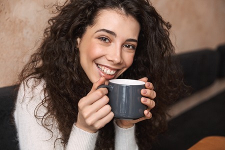 Image of pretty happy girl sitting in cafe drinking coffee holding cup.